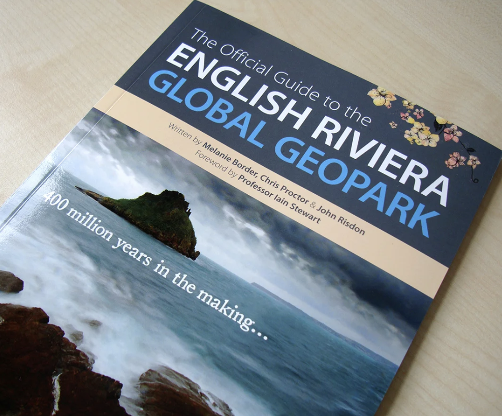 Book titled 'The Official Guide to the English Riviera and Global Geopark' resting on a wooden surface, with a cover image of a coastal scene featuring an island, ocean, and cloudy sky, and labeled with '400 million years in the making...'