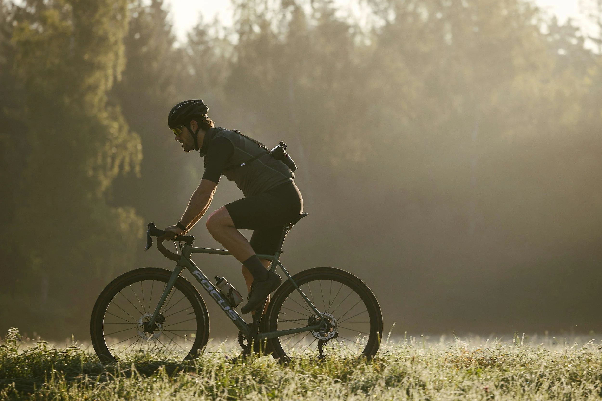 A man riding a bicycle through a grassy area at sunrise or sunset, wearing a helmet and sunglasses, with a blurred forest background.