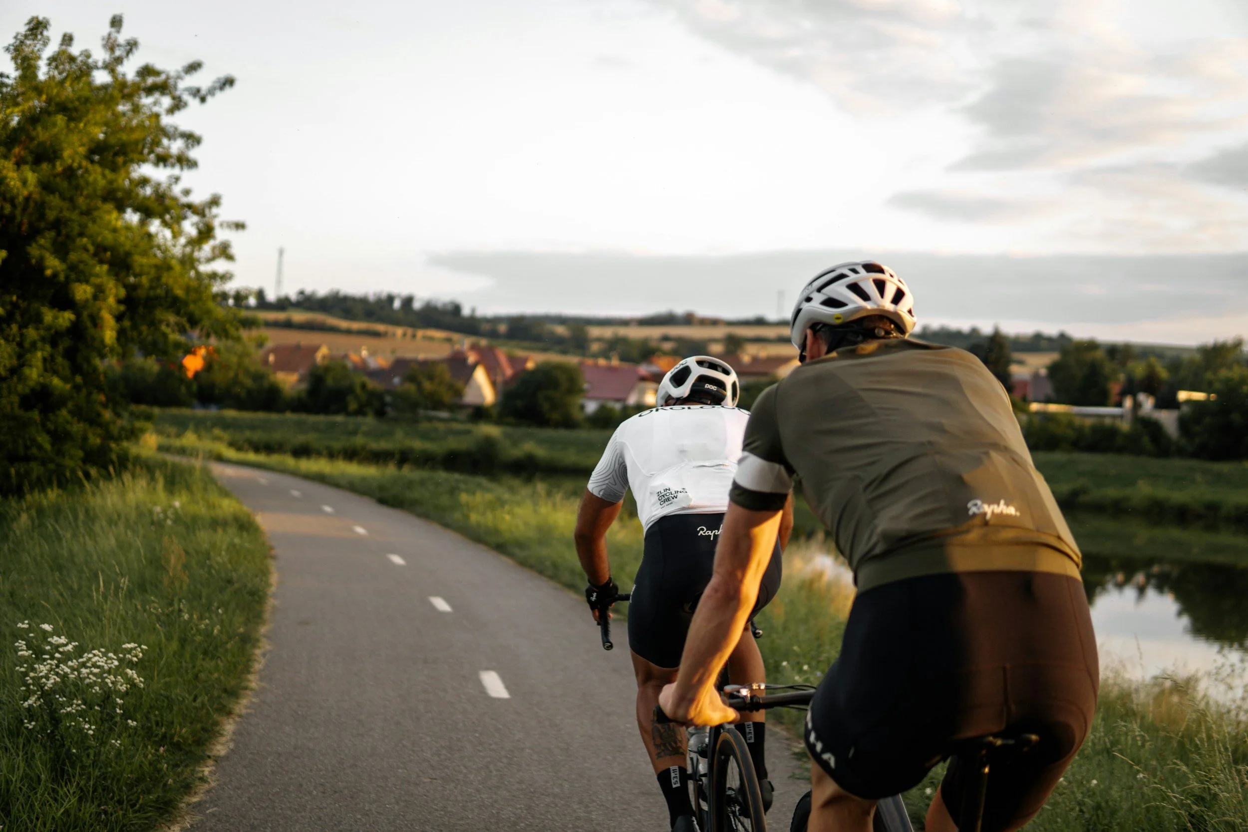 Two cyclists riding on a paved rural road with green grass and trees on the sides, houses and fields in the background, during sunset or early evening.