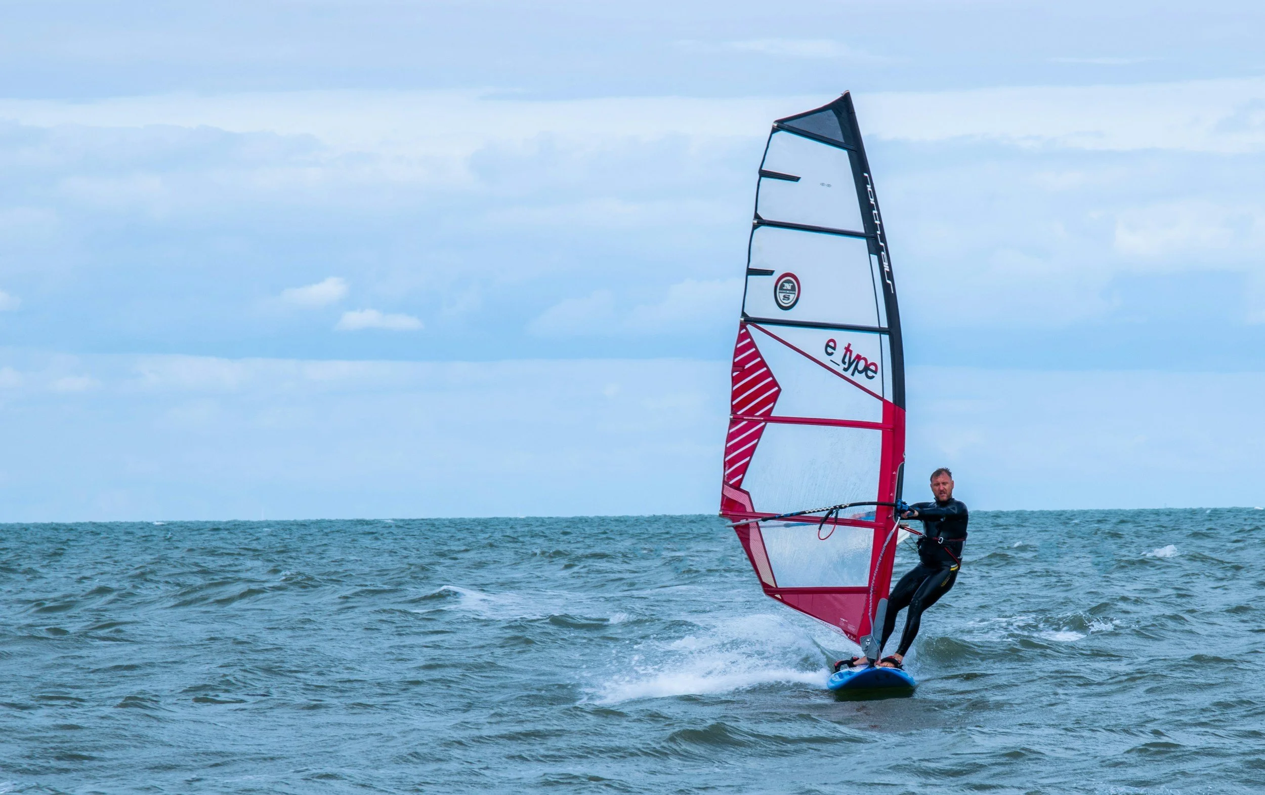A man windsurfing on the ocean with a sail labeled 'e-type' in the wind, under a partly cloudy sky.