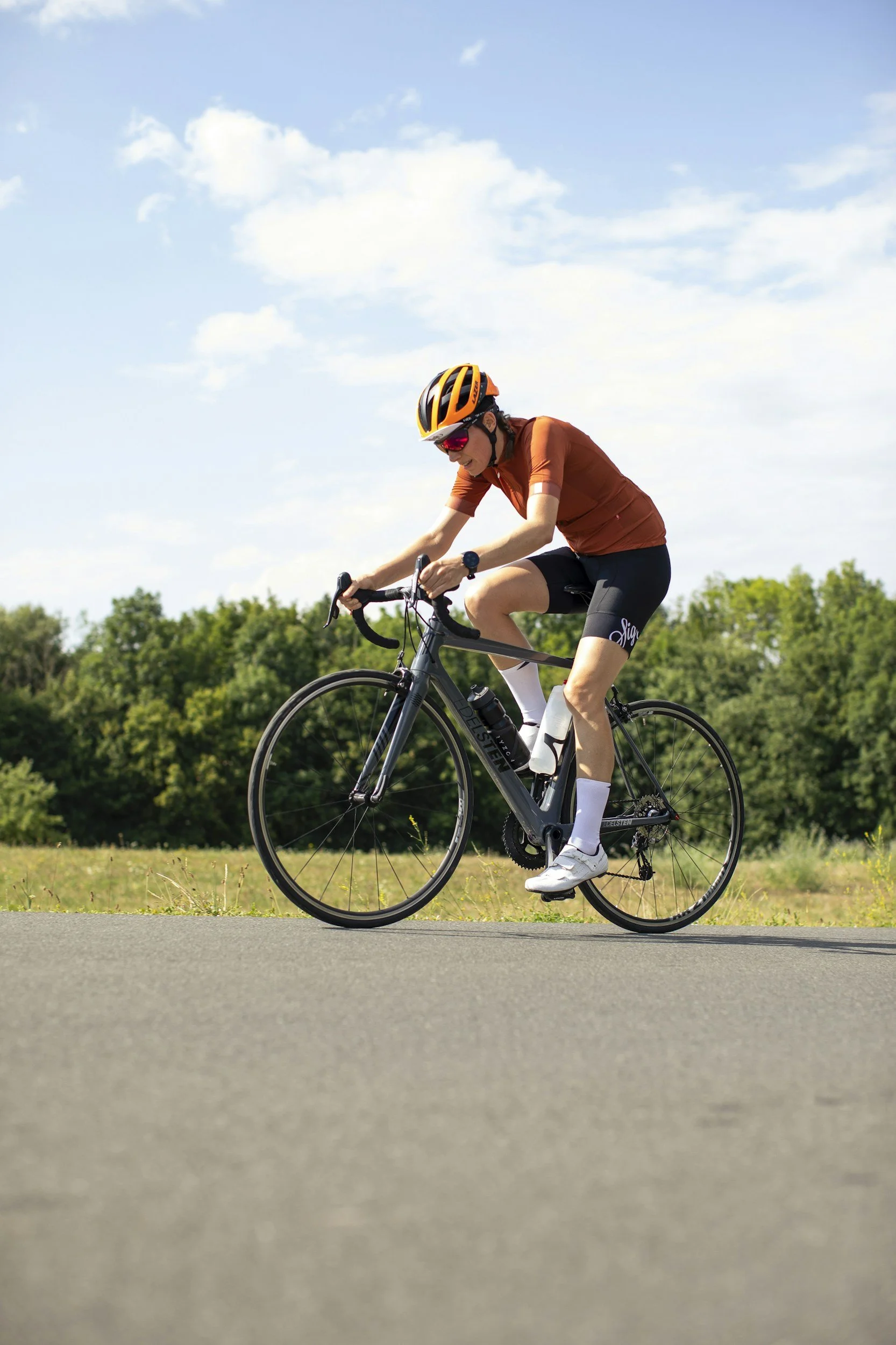 A person riding a bicycle on a paved path in a park, wearing an orange helmet and sunglasses, with trees and blue sky in the background.