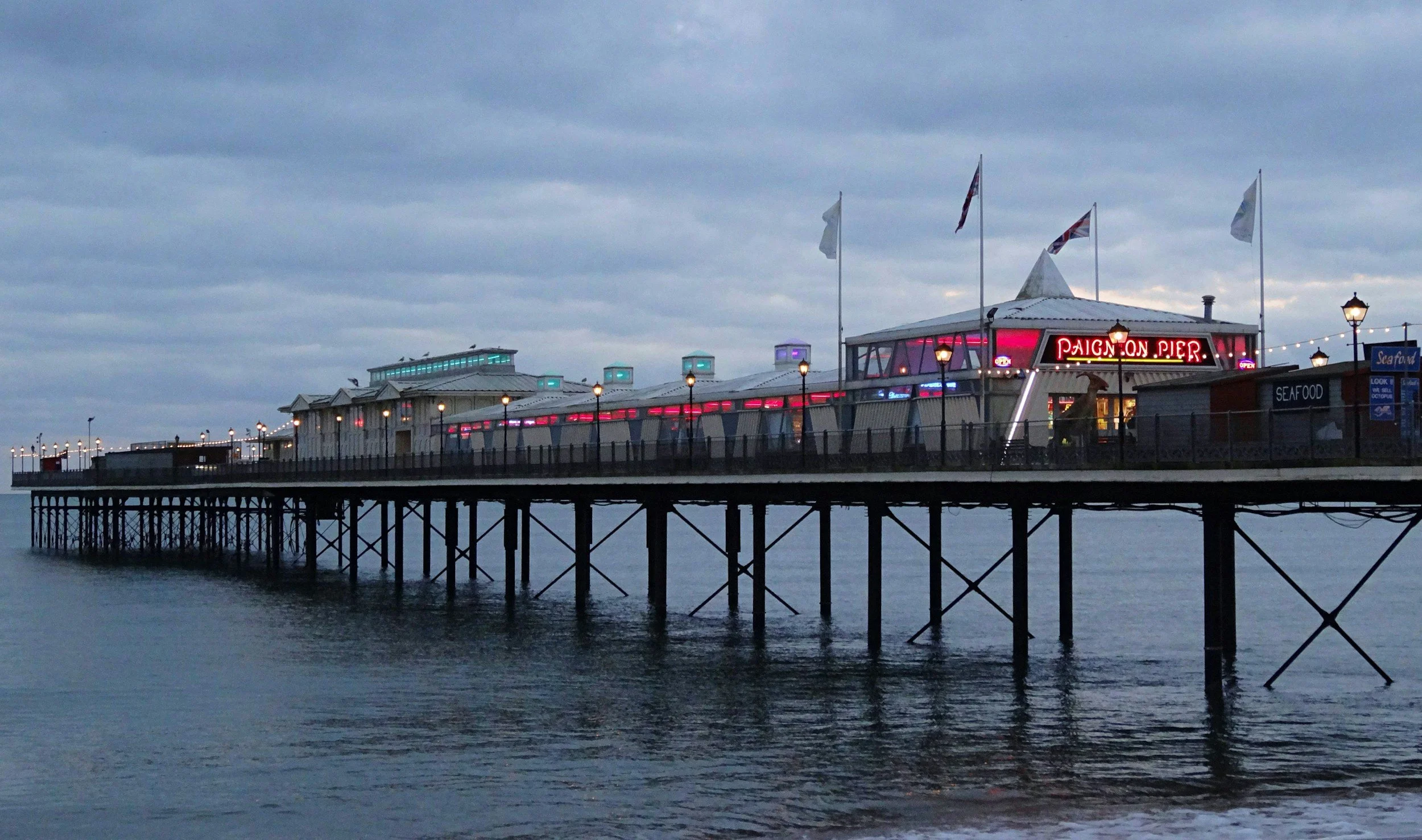 A pier over water with a building labeled 'Paimon Pier' illuminated with neon lights, flags flying on poles, and a cloudy sky overhead.