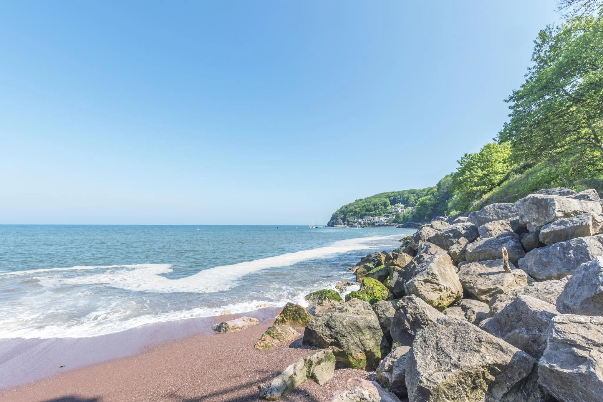 A beach with pinkish sand, large rocks along the shoreline, and calm ocean waters under a clear blue sky. Green trees and houses are visible on a hillside in the background.
