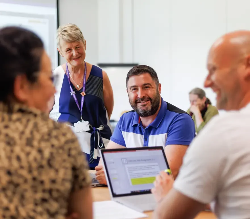 Four people in a meeting room engaged in discussion, smiling and looking at each other. One woman stands, wearing a blue sleeveless top, another woman is seated with her back to the camera, and two men are visible, one with a tablet and one with a laptop, all smiling.