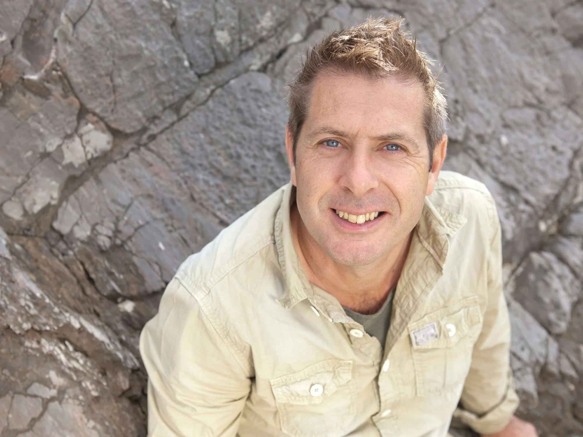 A smiling man with short, light brown hair and blue eyes, wearing a beige button-up shirt, standing outdoors in front of a rocky background.