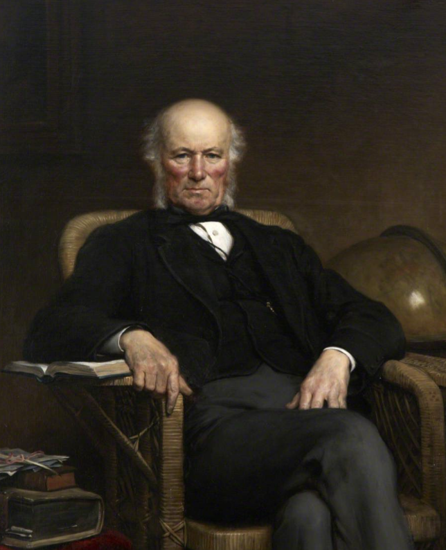 An elderly man with white hair, sitting in an armchair, wearing a black coat and vest, holding a book, with a globe behind him.