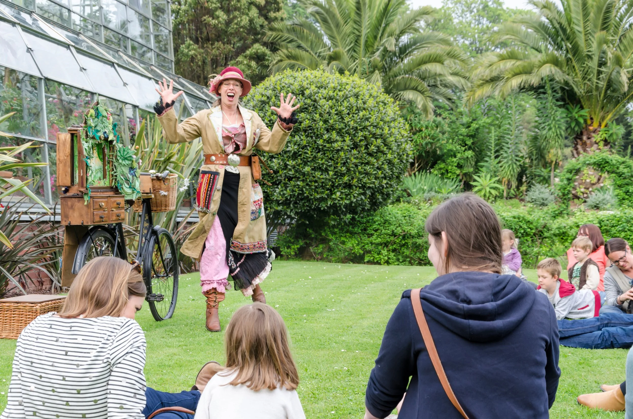 A woman dressed as a pirate performing for children sitting on the grass in a garden with lush green plants and trees.
