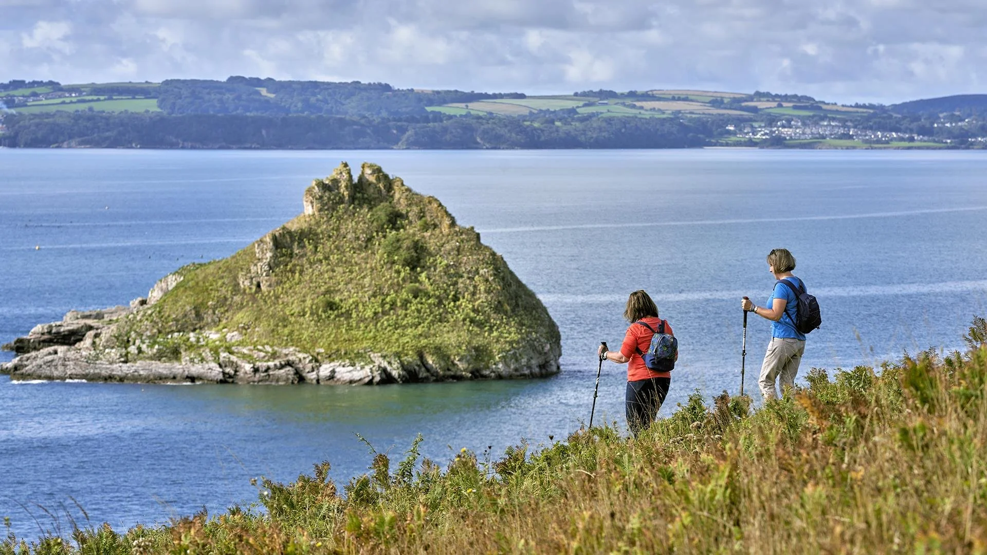 Two hikers with walking poles and backpacks walk along a grassy hill overlooking the ocean with a rocky, moss-covered island in the water. In the background, there are rolling hills with patches of green farmland and a distant shoreline.