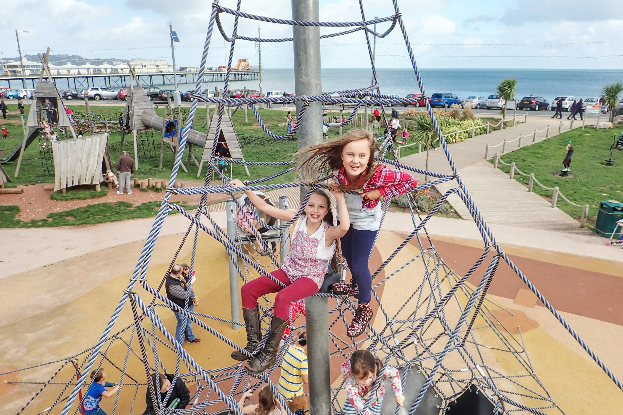 Two young girls playing on a rope climbing structure at an outdoor playground near the beach, with the ocean and a pier in the background.