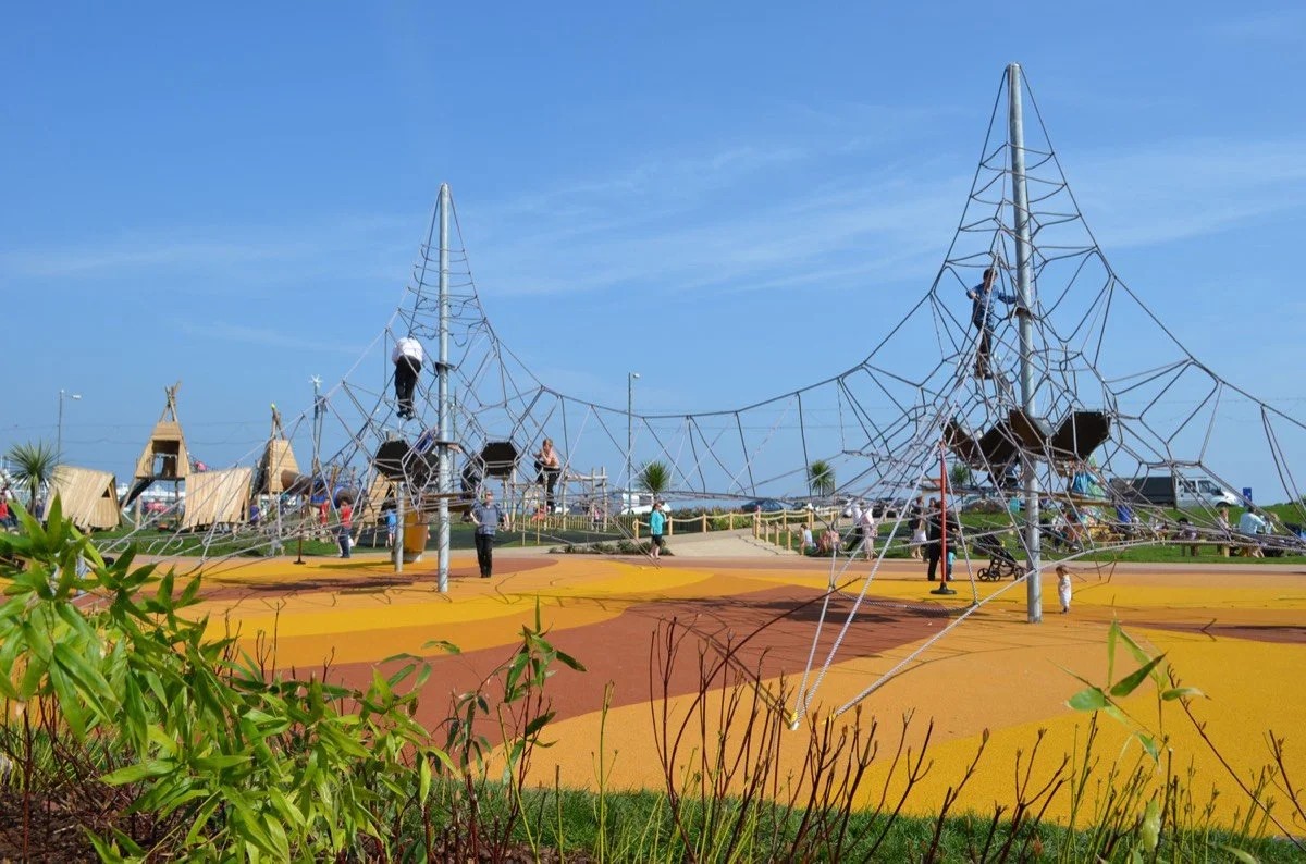 Children climbing on a large spider web playground structure at a park with blue sky and a colorful ground surface.