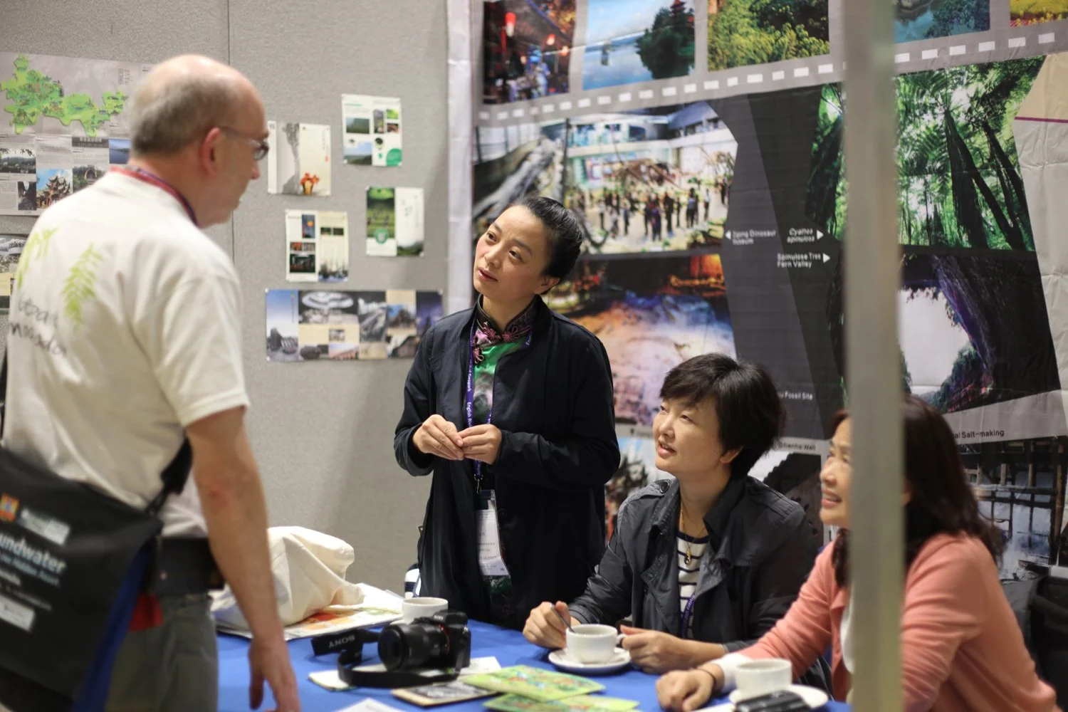 A group of four people having a conversation at a booth, with two women sitting at a table with coffee cups and a camera, and a man standing and talking to them, in front of a display of nature photographs.