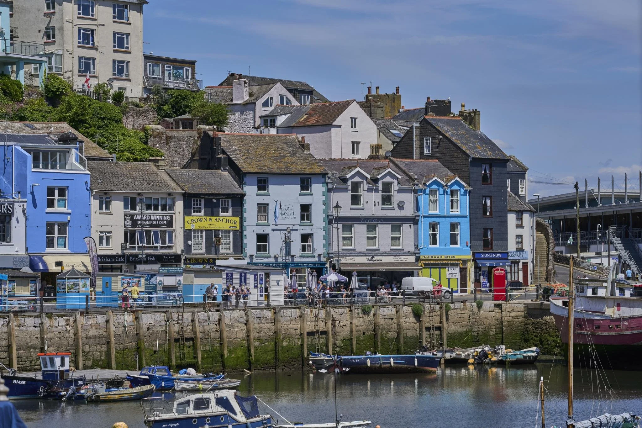 A seaside town with colorful buildings along the waterfront, boats docked in the harbor, people walking by the water, and hillside houses in the background under a partly cloudy sky.