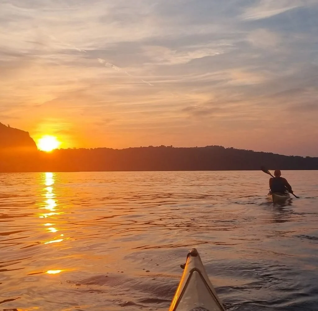 A person kayaking on a body of water during a sunset, with a scenic view of the horizon and colorful sky.
