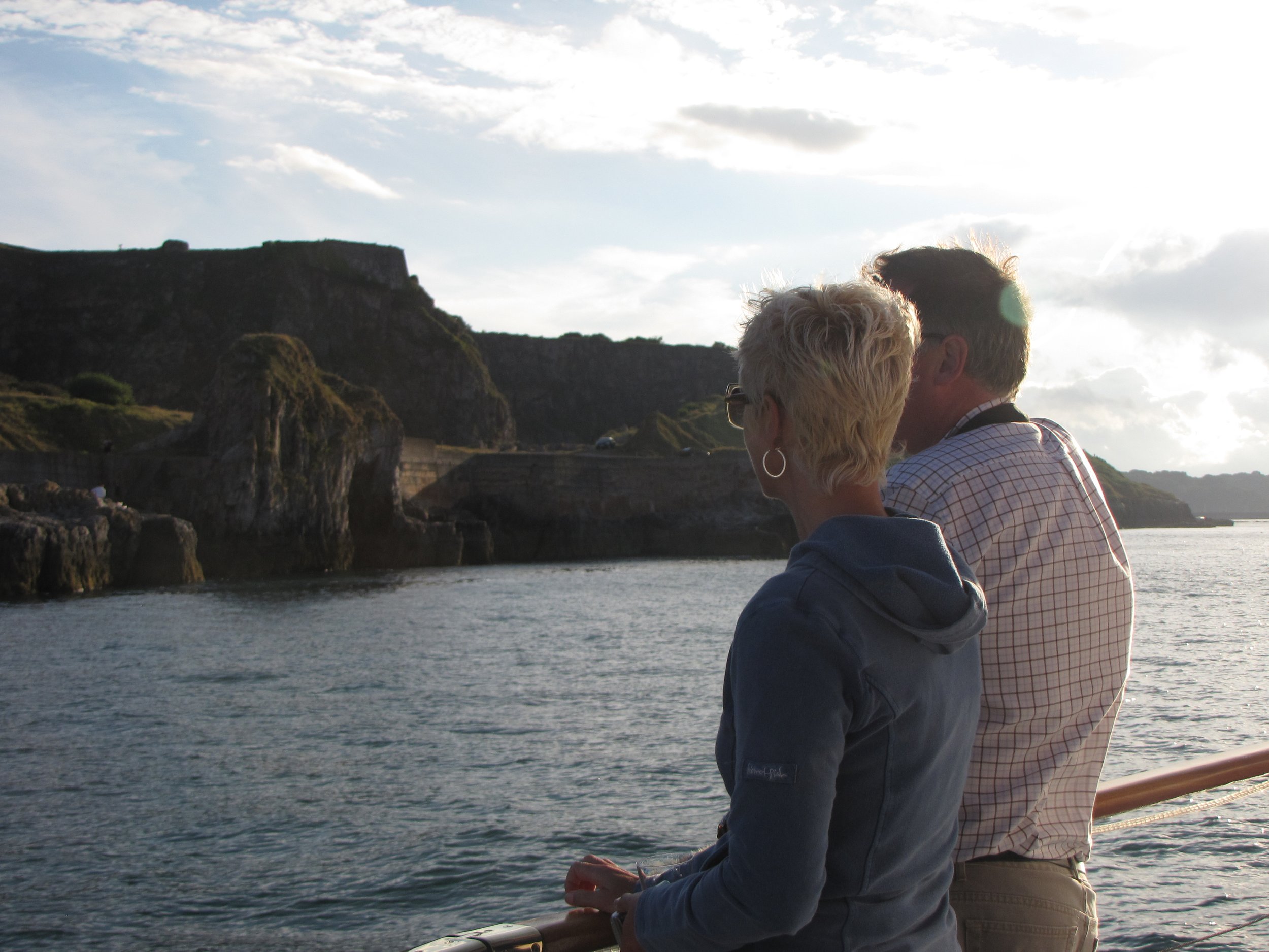 Two people stand on a boat or dock, looking at a coastal landscape with cliffs, rocky formations, and water. The sky is partly cloudy with sunlight shining through.