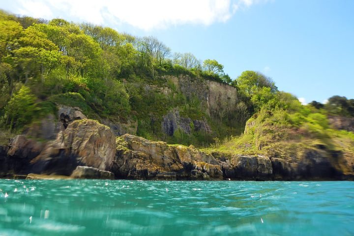 Waterfront view with turquoise water, rocky shoreline, and green forested cliffs under a partly cloudy sky.