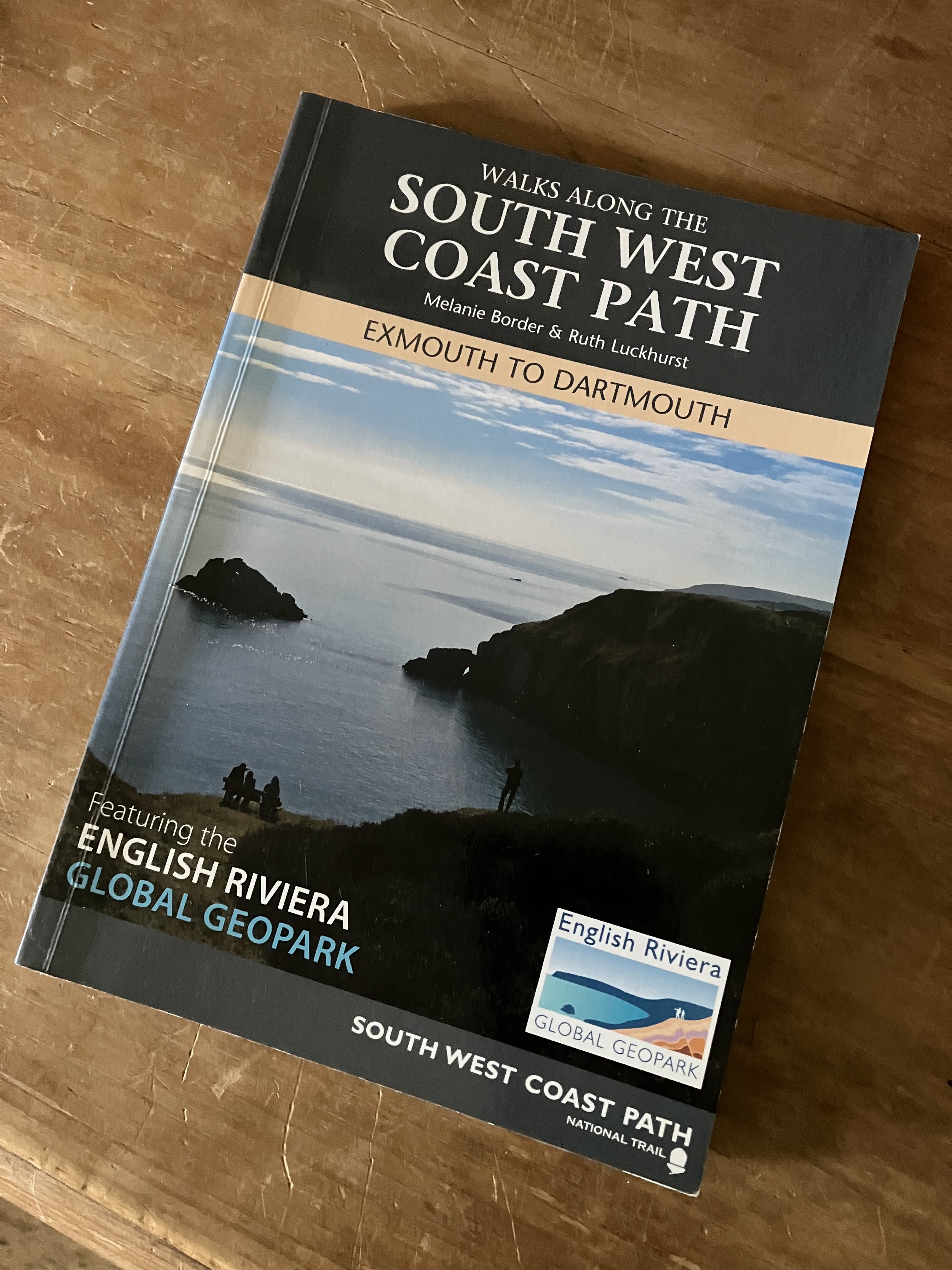 A guidebook titled "Walks Along the South West Coast Path" by Melanie Border and Ruth Luckhurst, featuring a coastal landscape with rocky cliffs and the sea at sunset.
