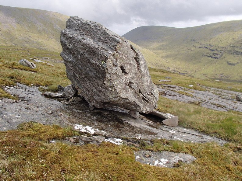 Large tilted boulder resting on smaller rocks in a grassy, hilly landscape with cloudy sky.