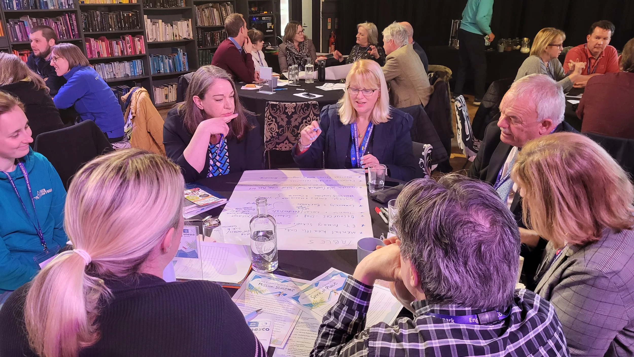 Group of people seated around tables engaged in discussion at a workshop or conference in a library or meeting room, with bookshelves in the background.