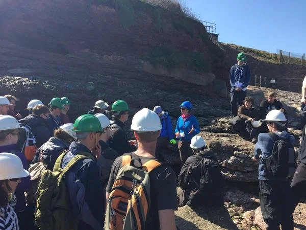 Group of students wearing helmets and backpacks on a geology field trip, listening to a guide in a rocky outdoor area.