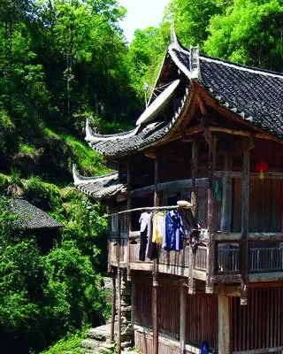 Traditional wooden house with curved roof, balconies, and hanging clothes surrounded by lush green trees.