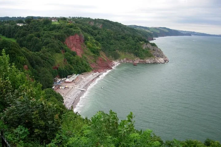 A scenic view of a coastline with a beach, green trees on the cliffs, and a body of water under a cloudy sky.