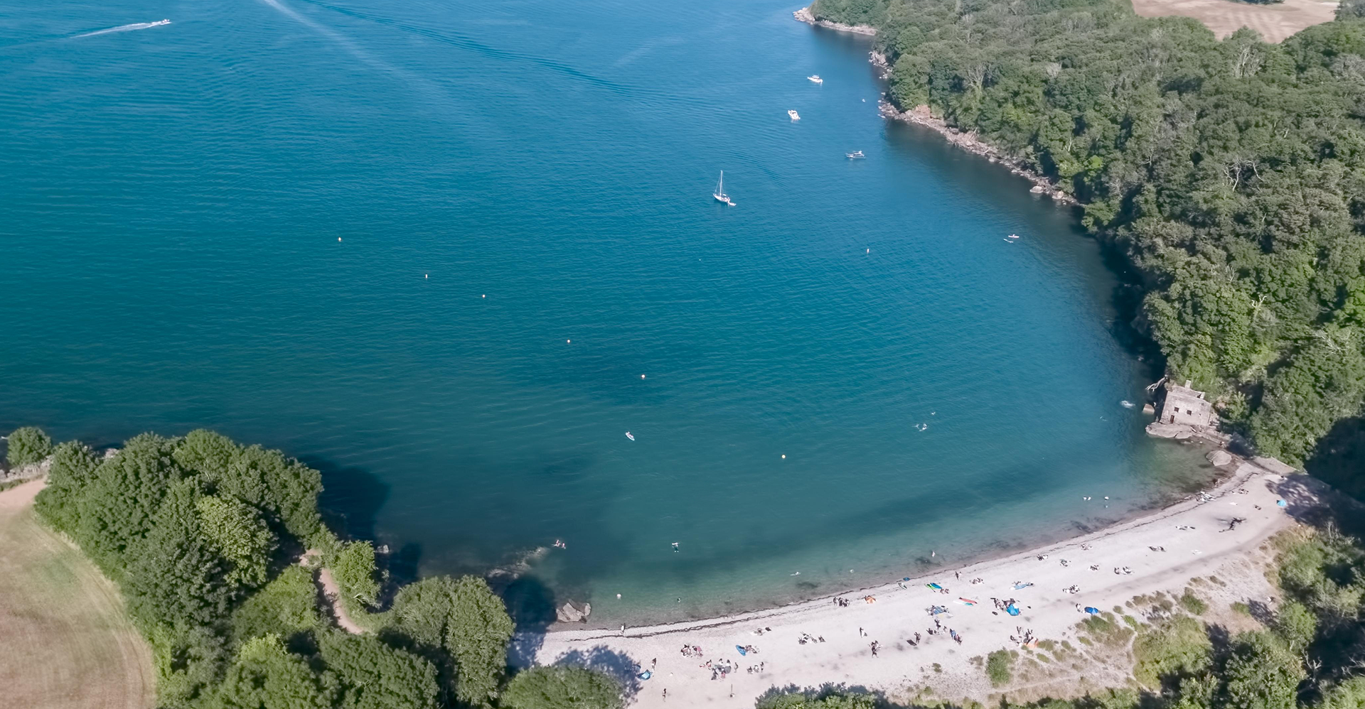 Aerial view of a small sandy beach with colorful umbrellas and people, surrounded by green trees and water with boats, near a wooded shoreline.