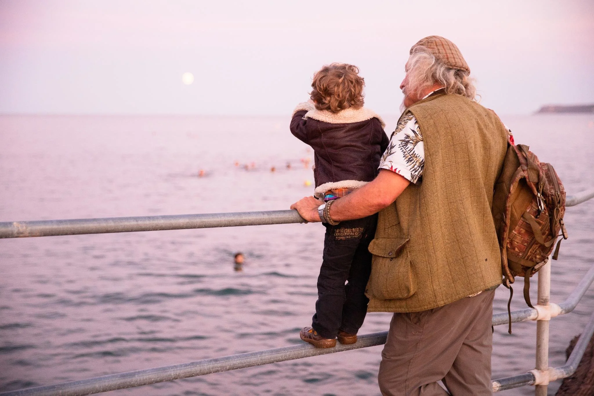An elderly man with gray hair wearing a beige vest and a patterned shirt, holding a child with curly hair, at the railing of a waterfront during sunset, with a pinkish sky and calm water in the background.