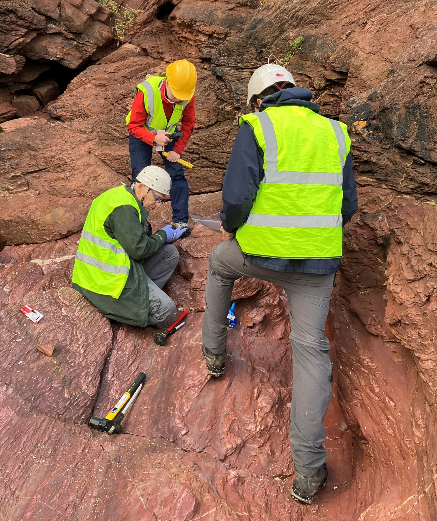 Four geology researchers wearing yellow safety vests, helmets, and outdoor gear examining reddish rock formations and using hand tools in a cliff face or canyon environment.