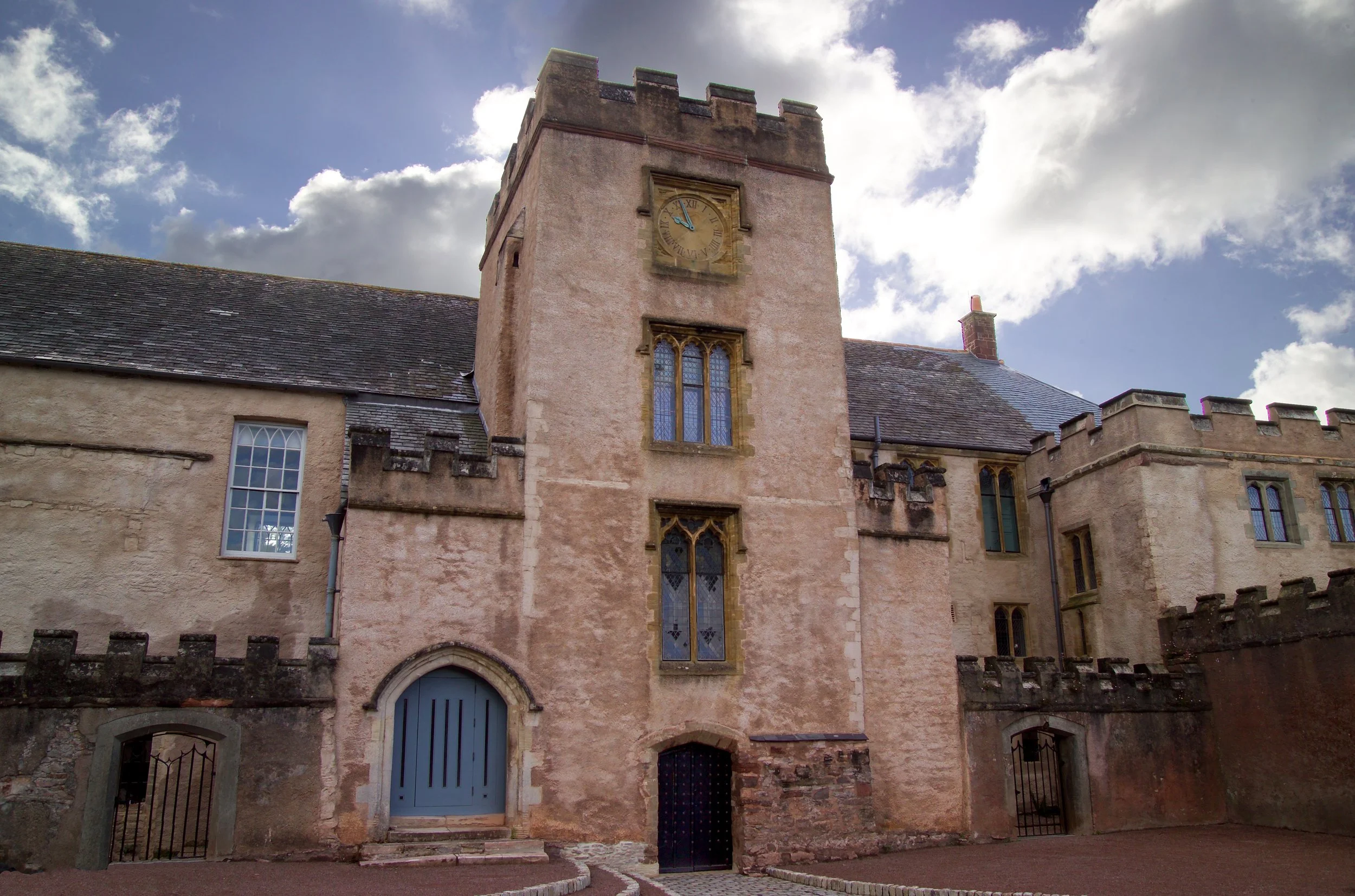 A historic stone building with a clock tower, featuring Gothic-style windows and a crenellated parapet, under a partly cloudy sky.