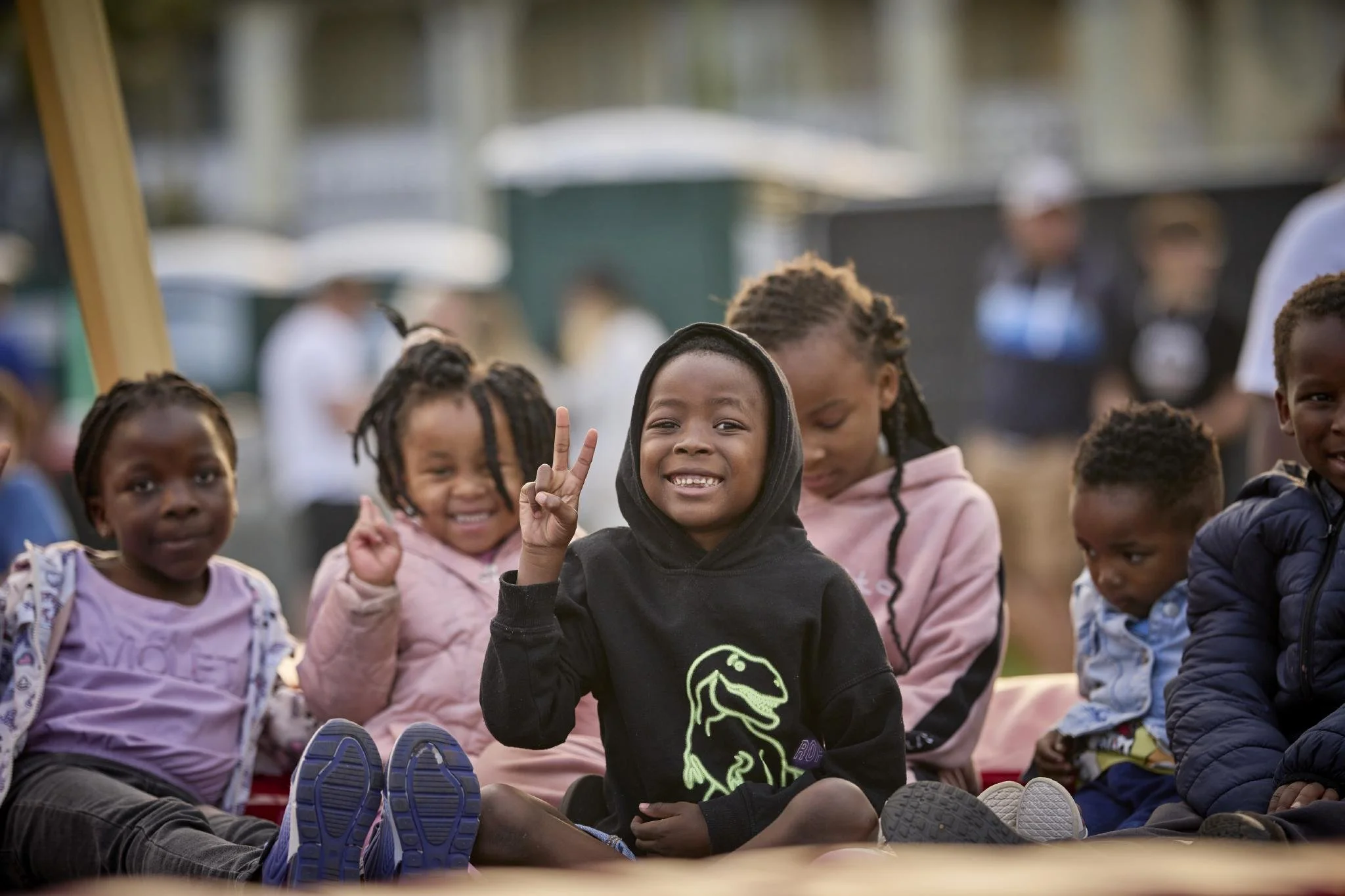 Children sitting outdoors, smiling and making peace signs, with some wearing hoodies and jackets. Background shows tents or temporary structures and blurred people.
