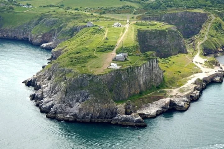 A cliffside with green grass, small buildings, and pathways overlooking the ocean.