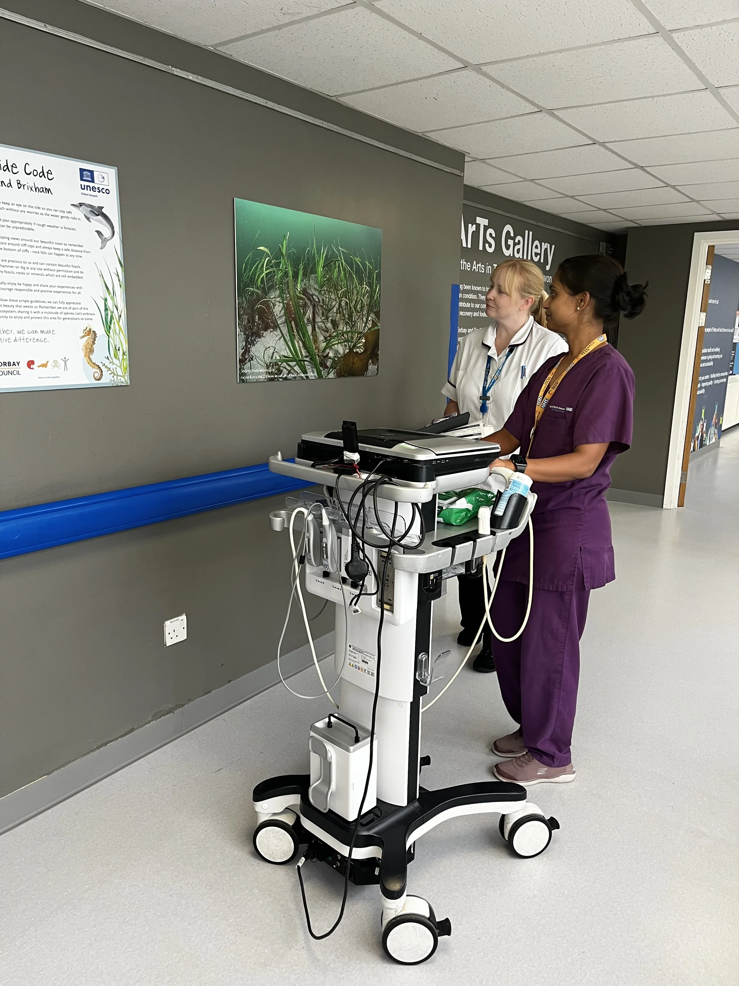 Two healthcare workers in scrubs working with medical equipment on a mobile cart in a hospital corridor, with informational posters on the wall.