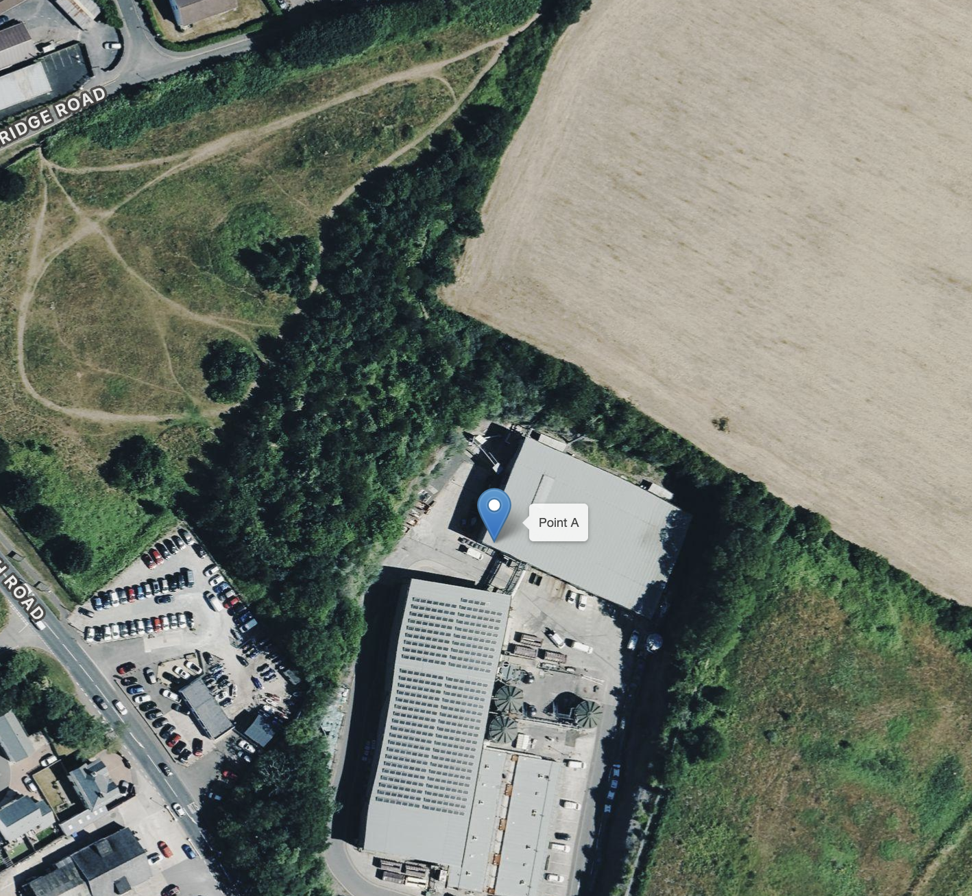 An aerial view of a parking lot filled with cars, next to a large industrial building, surrounded by trees, fields, and a road labeled 'Bridge Road.' A marker labeled 'Point A' is on the building.