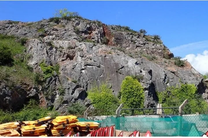 An outdoor swimming pool with a green fence in front of a rocky hillside with trees and bushes.