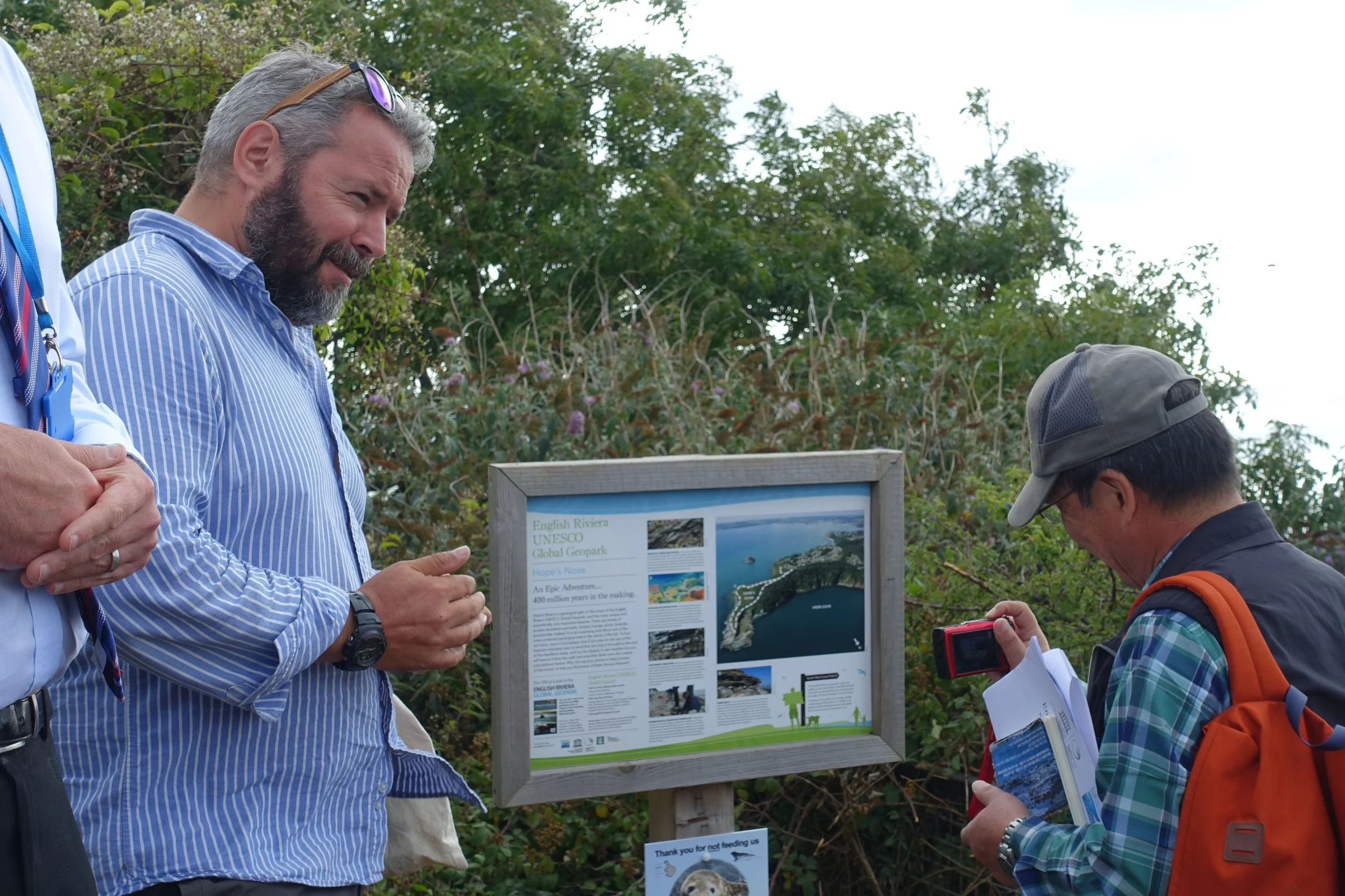 Two men are standing outdoors near an informational sign about the English Riviera UNESCO Global Geopark. One man, with gray hair and a beard, is pointing at the display while the other, wearing a cap and backpack, is holding a phone. The background features green trees and shrubs.