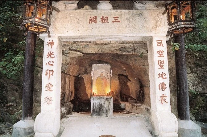 A shrine with a stone statue of a seated figure, possibly a Buddhist figure, inside a cave. The shrine is framed by white stone pillars with Chinese characters and decorative lanterns hanging on both sides.