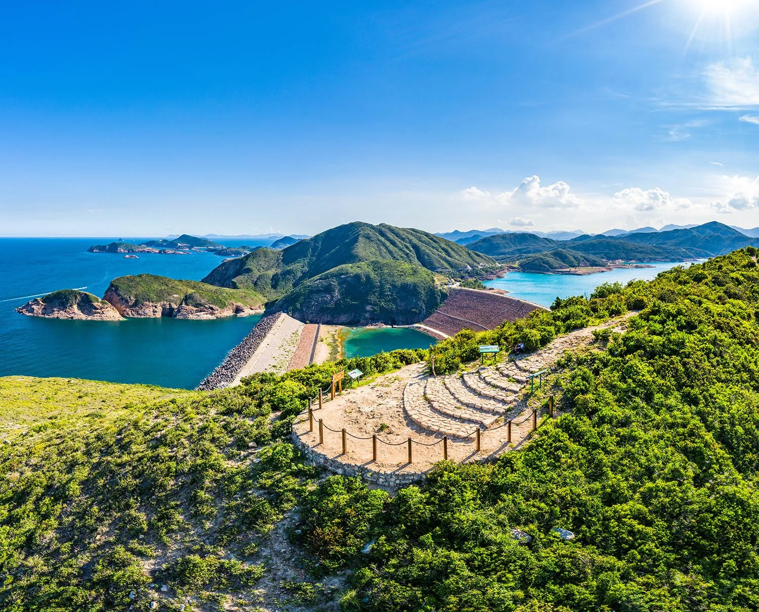 Scenic view of a dam with water reservoirs, surrounded by green mountains, under a clear blue sky with the sun shining.