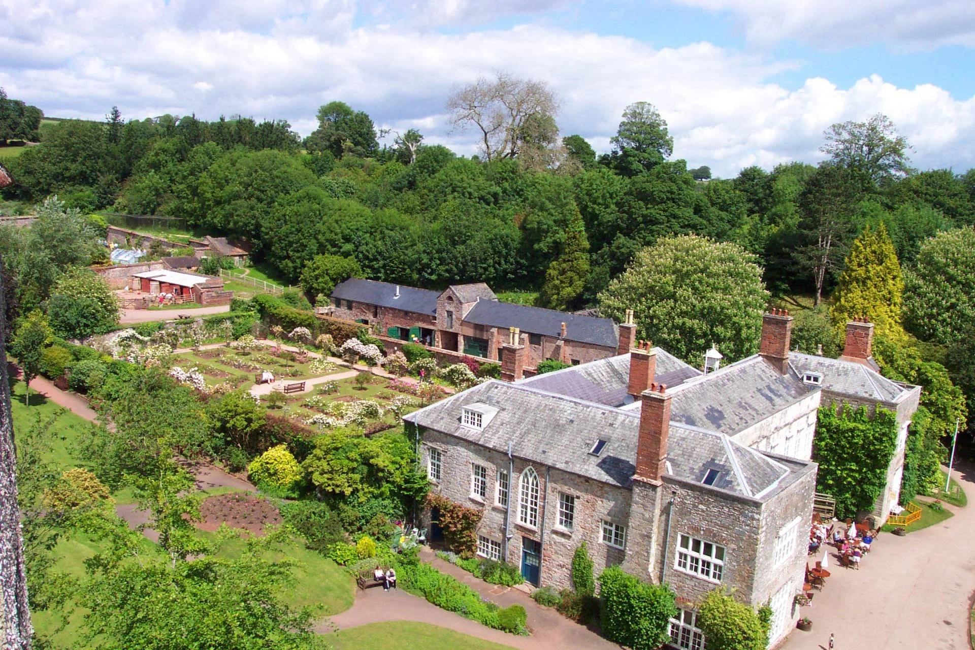 A large historic stone building with multiple chimneys and a slate roof, surrounded by a manicured garden with paths, benches, and blooming flowers. In the background, there is a lush green forest under a partly cloudy sky.