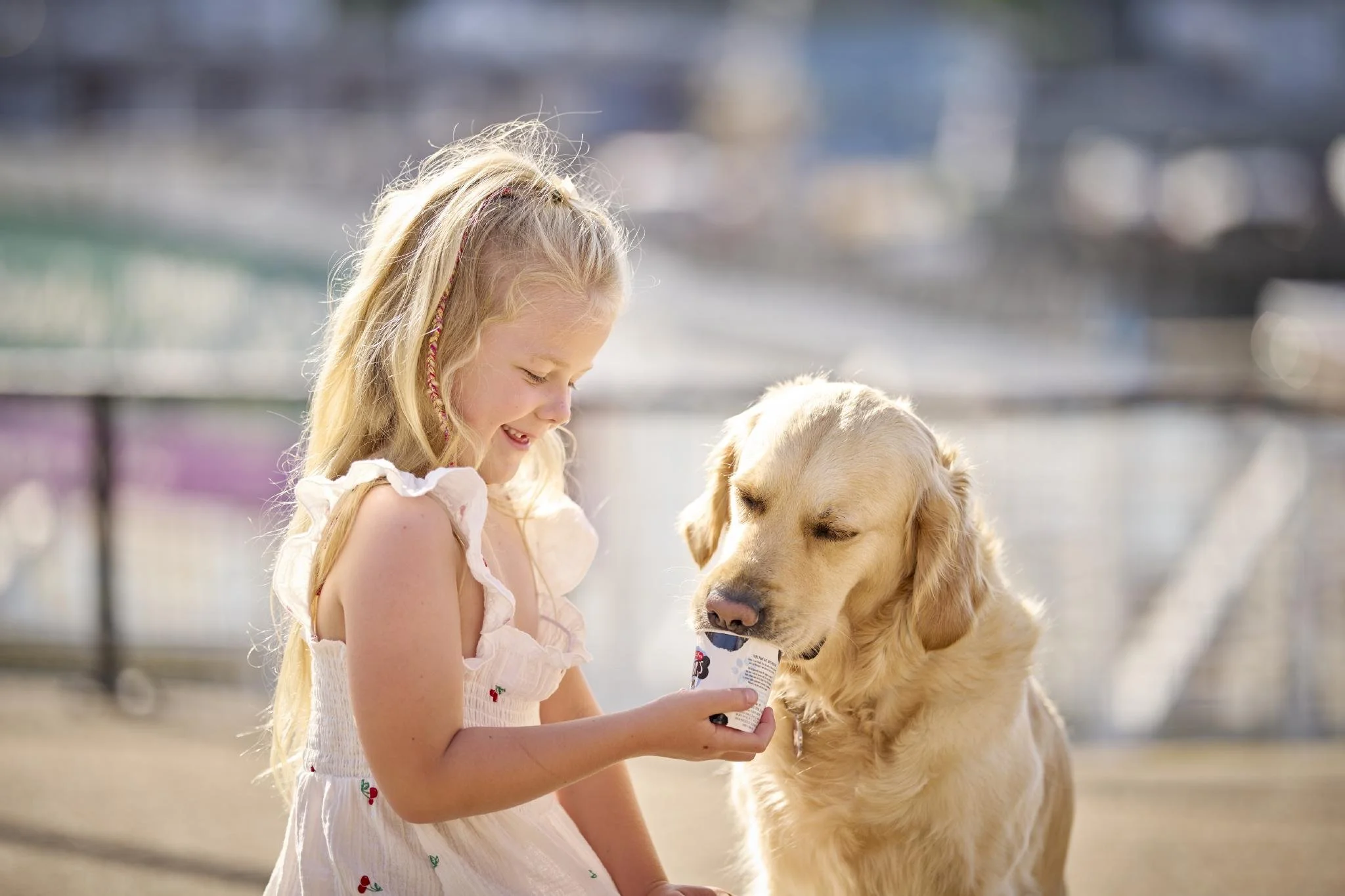 A young girl with long blonde hair and a light dress is smiling and holding a cup of ice cream while a golden retriever dog with closed eyes is sitting beside her on a sunny day outdoors.