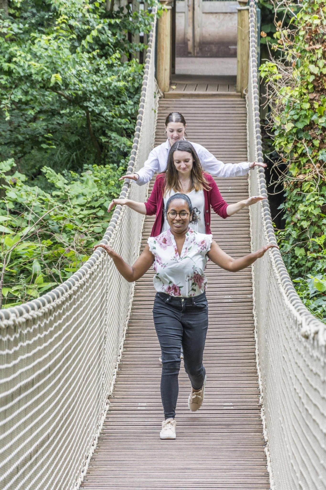 Four women walking in a single file across a suspension bridge surrounded by greenery, with arms extended to balance.