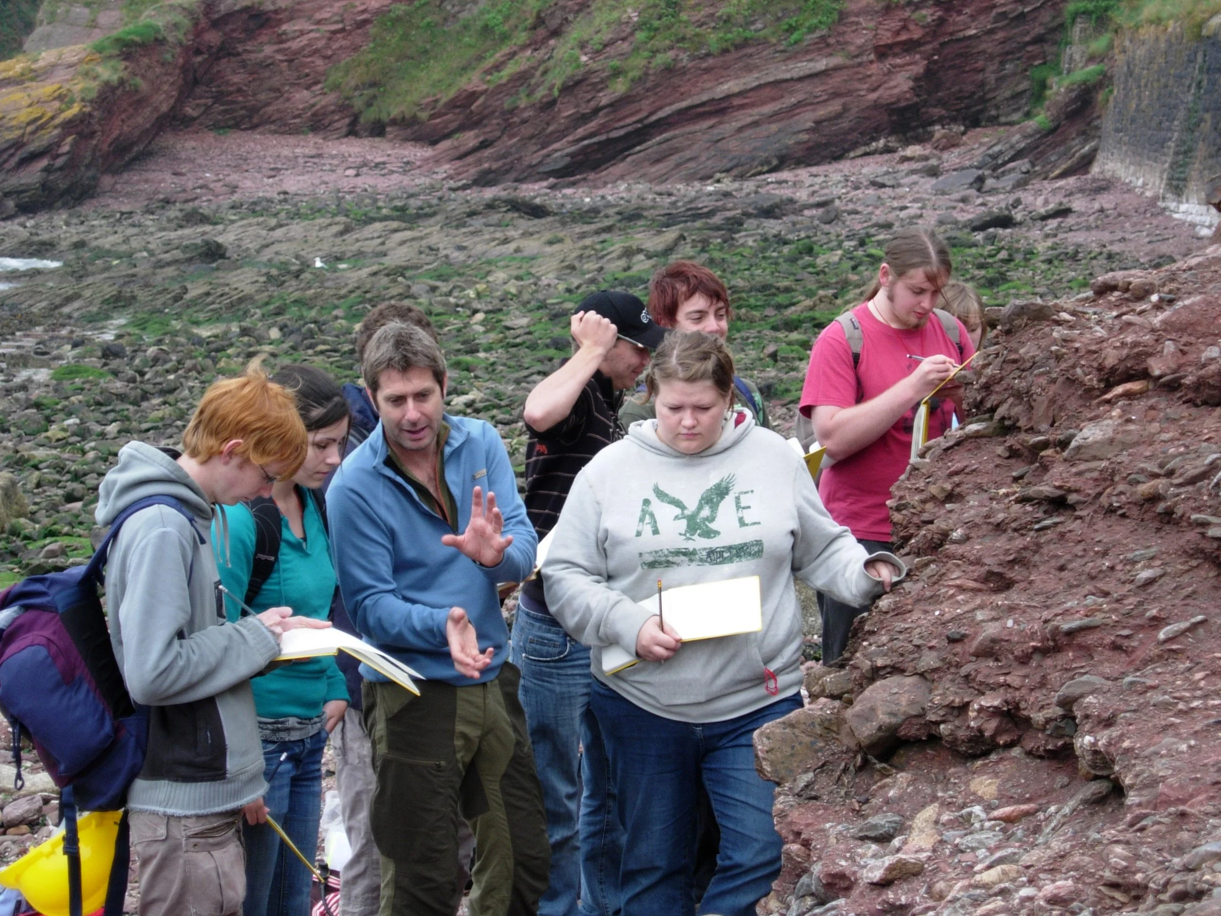 Group of people gathered outdoors at a rocky cliff, studying or observing the rocks closely, some holding notebooks and pens.