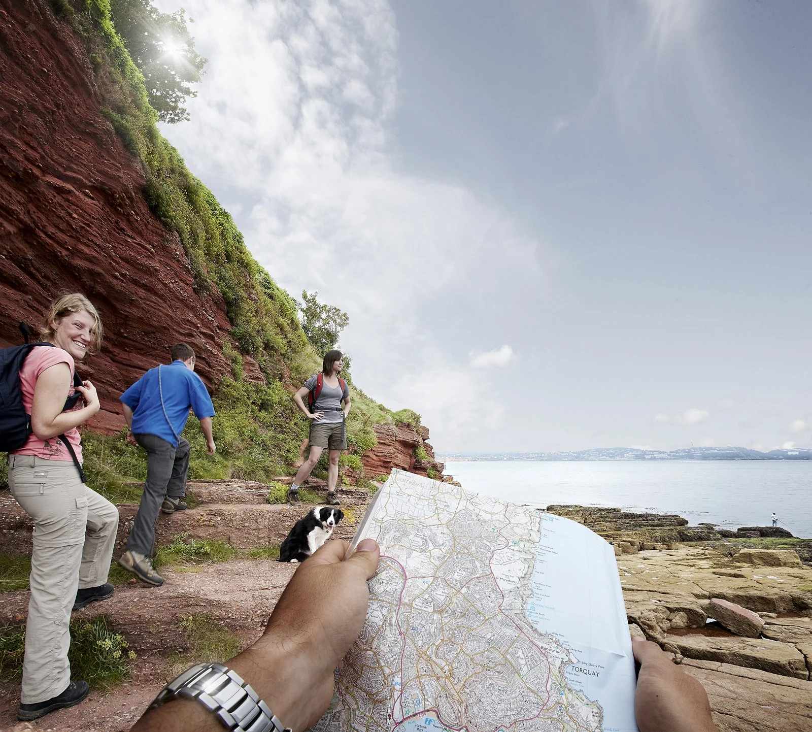 A group of hikers on a coastal trail, with a red cliff and ocean in the background. One person holds a map, and a dog sits near them. The scene is sunny with a partly cloudy sky.
