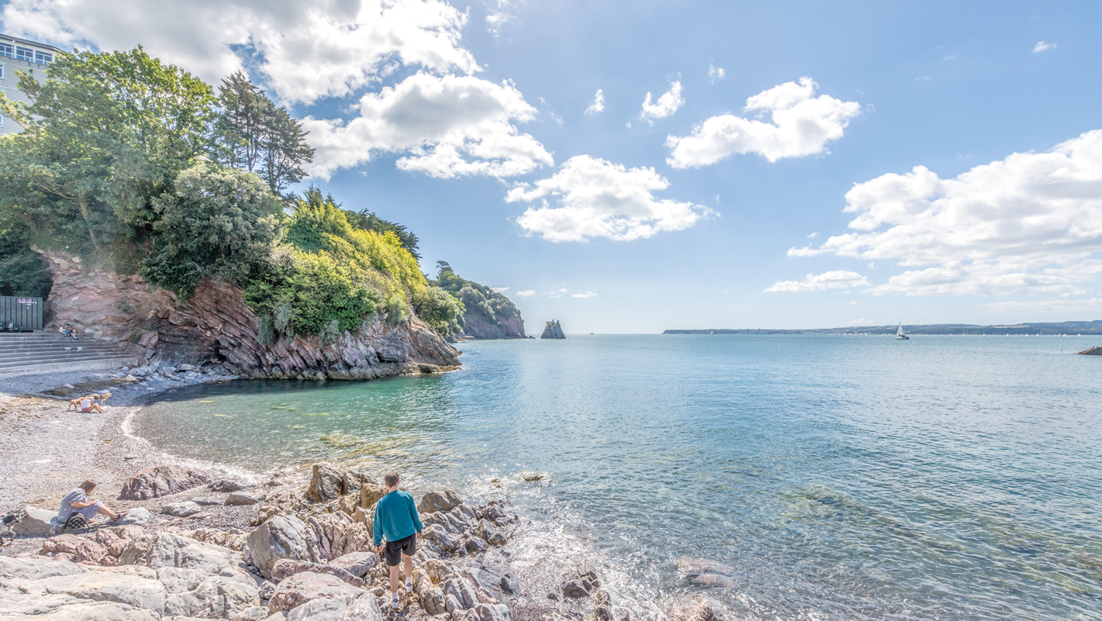 Beach with rocks, trees, and a few people relaxing, sailing boat on calm water, partly cloudy sky.