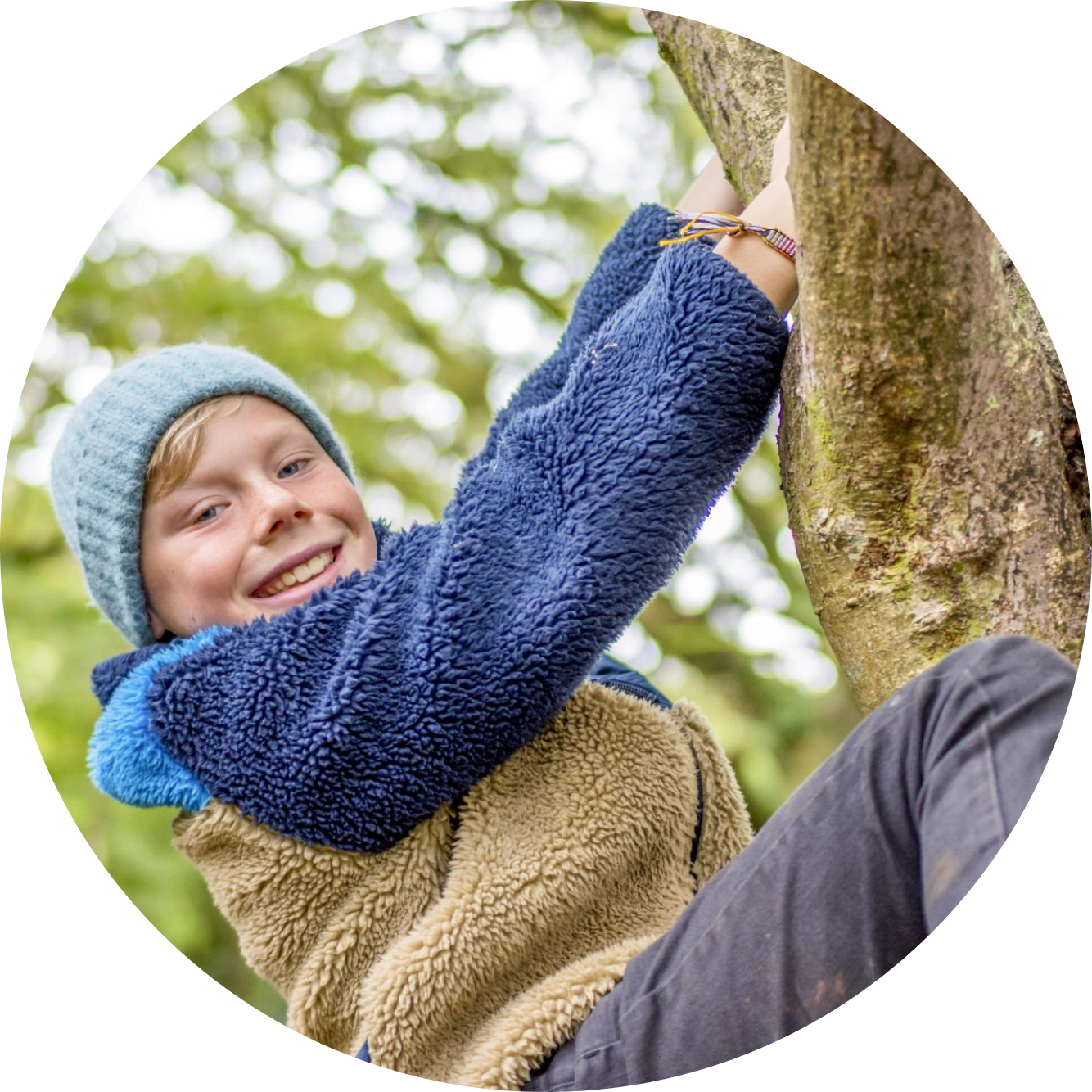 A young boy wearing a blue knit hat and a fleece jacket is smiling while climbing a tree outdoors.