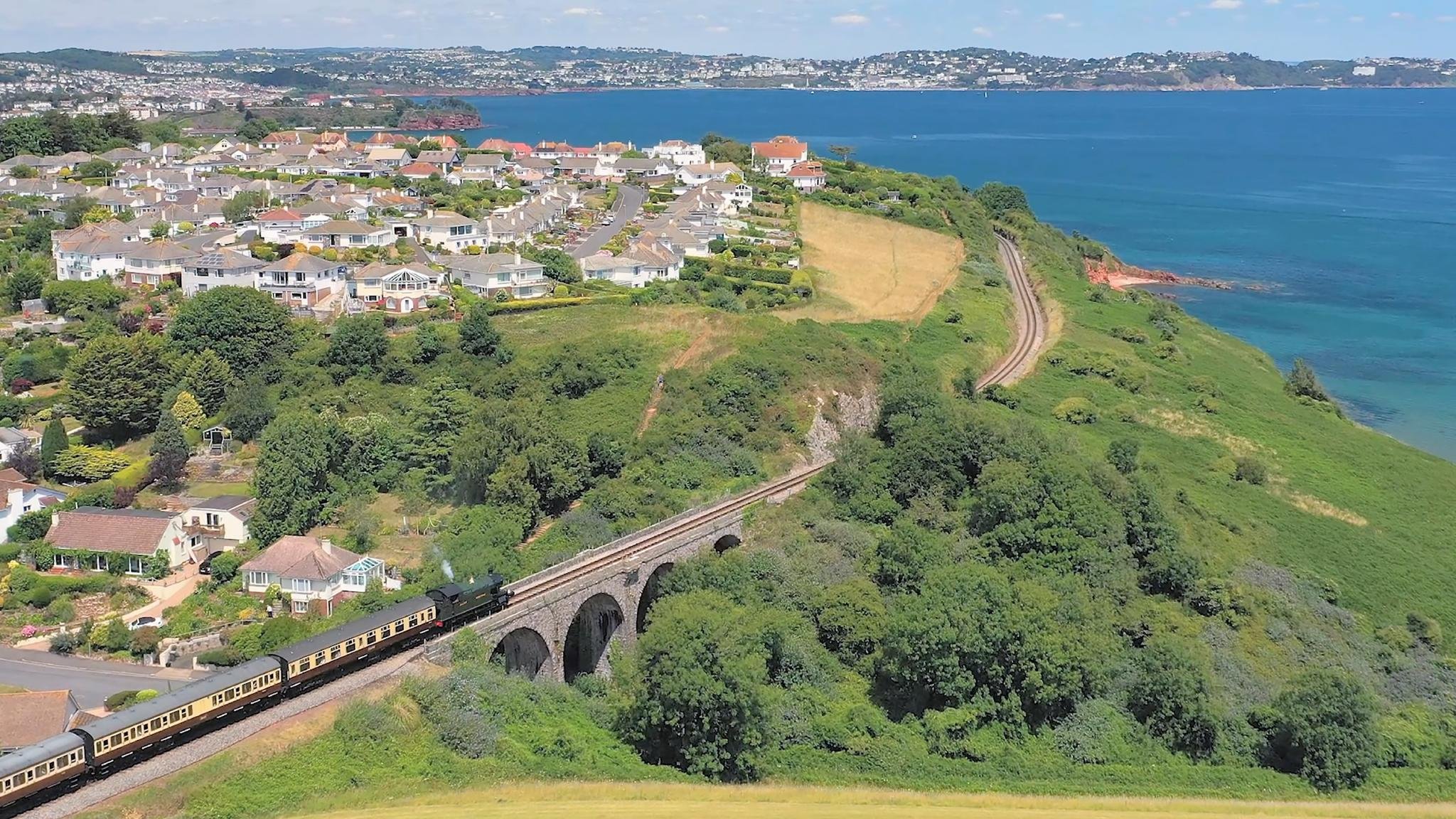 A scenic view of a suburban neighborhood near a coast with houses, greenery, and a train crossing a stone bridge.