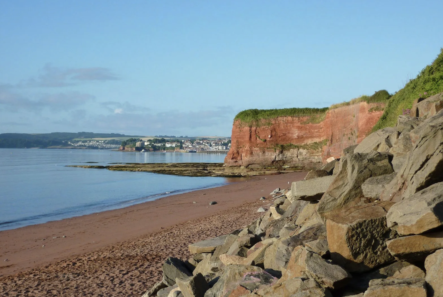 Cliffs and sandy beach along the shoreline with calm water and a distant town in the background under a clear blue sky.