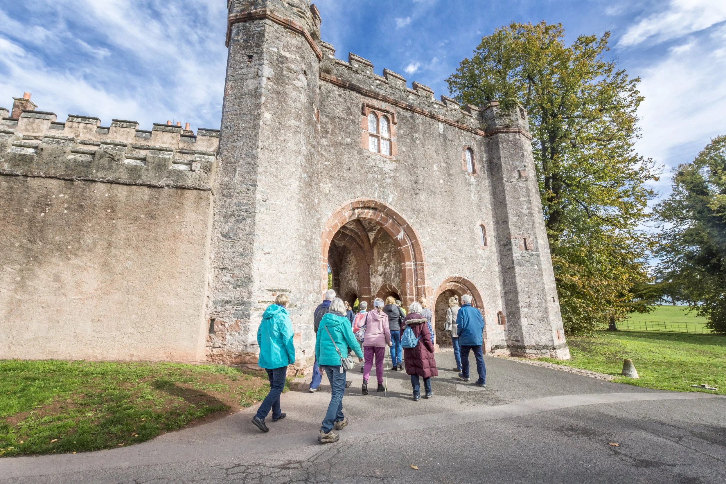 A group of tour visitors walking towards the entrance of a historic stone castle with tall walls, arched doorway, and small windows, surrounded by green trees under a partly cloudy sky.