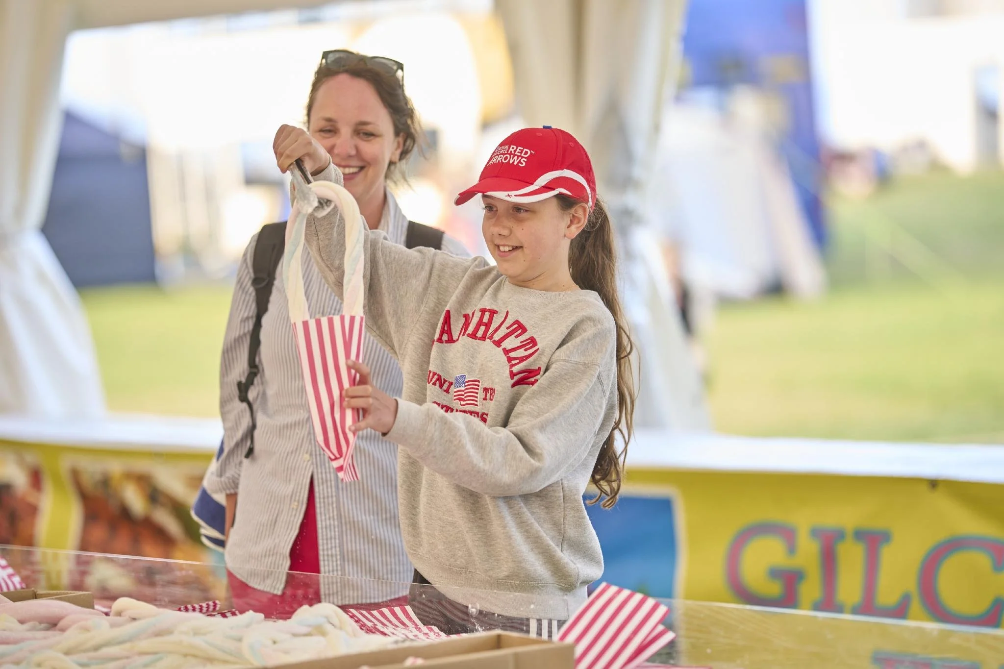 A woman and a girl at an outdoor fair examining pink and white twisted candies in a striped paper bag.