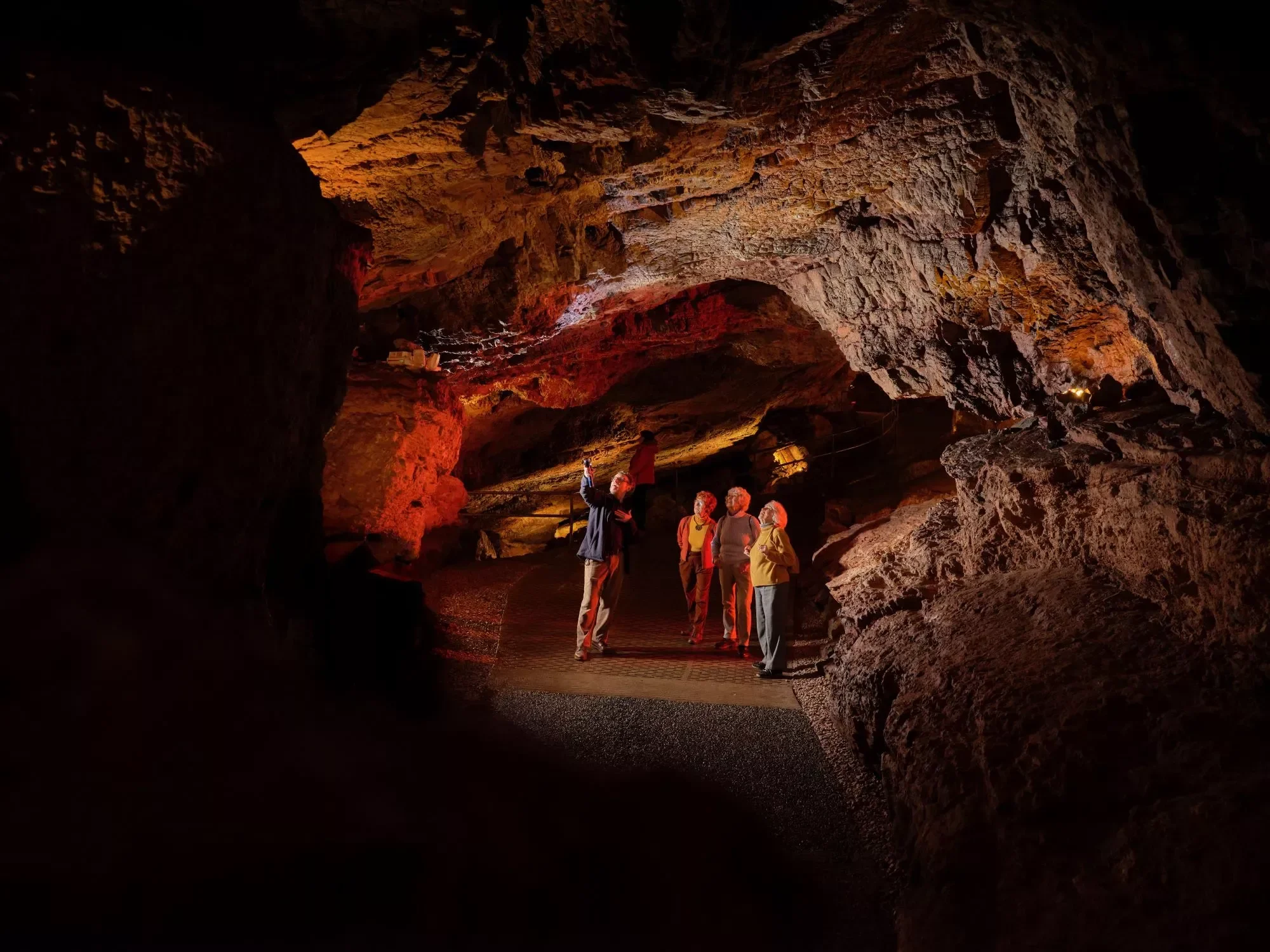 A group of people inside a large, rocky cave lit with warm, orange lighting, some of whom are taking photographs or looking around.