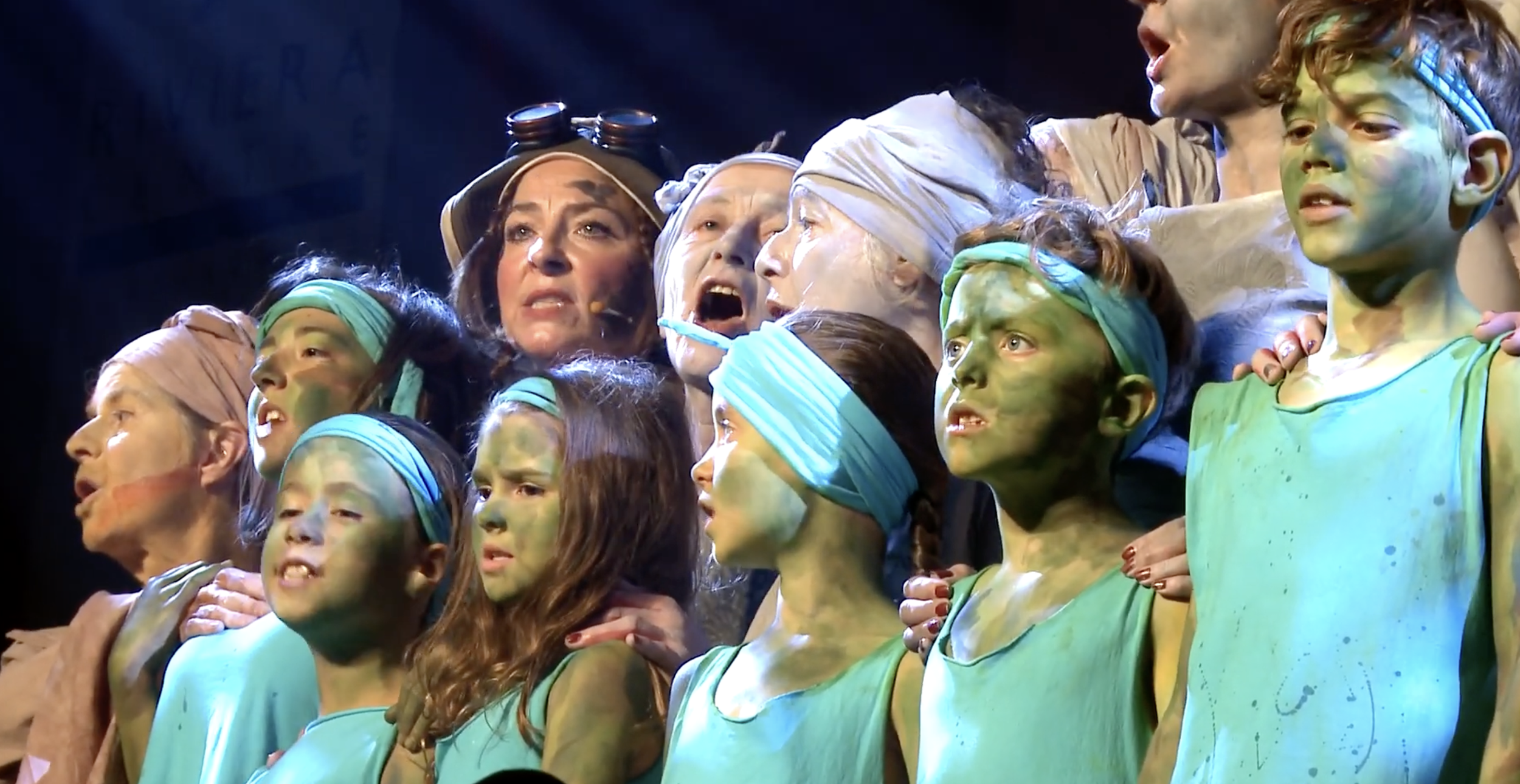 Group of people on stage, with green and blue face paint, some with headbands or goggles, appearing to sing or perform in a theatrical production.